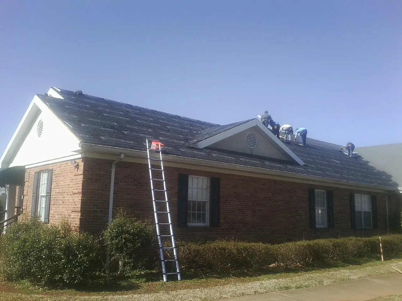 Crew tearing shingles off a brick ranch roof with a ladder set — Emergency Roof Repair work by our crew in East Lansing