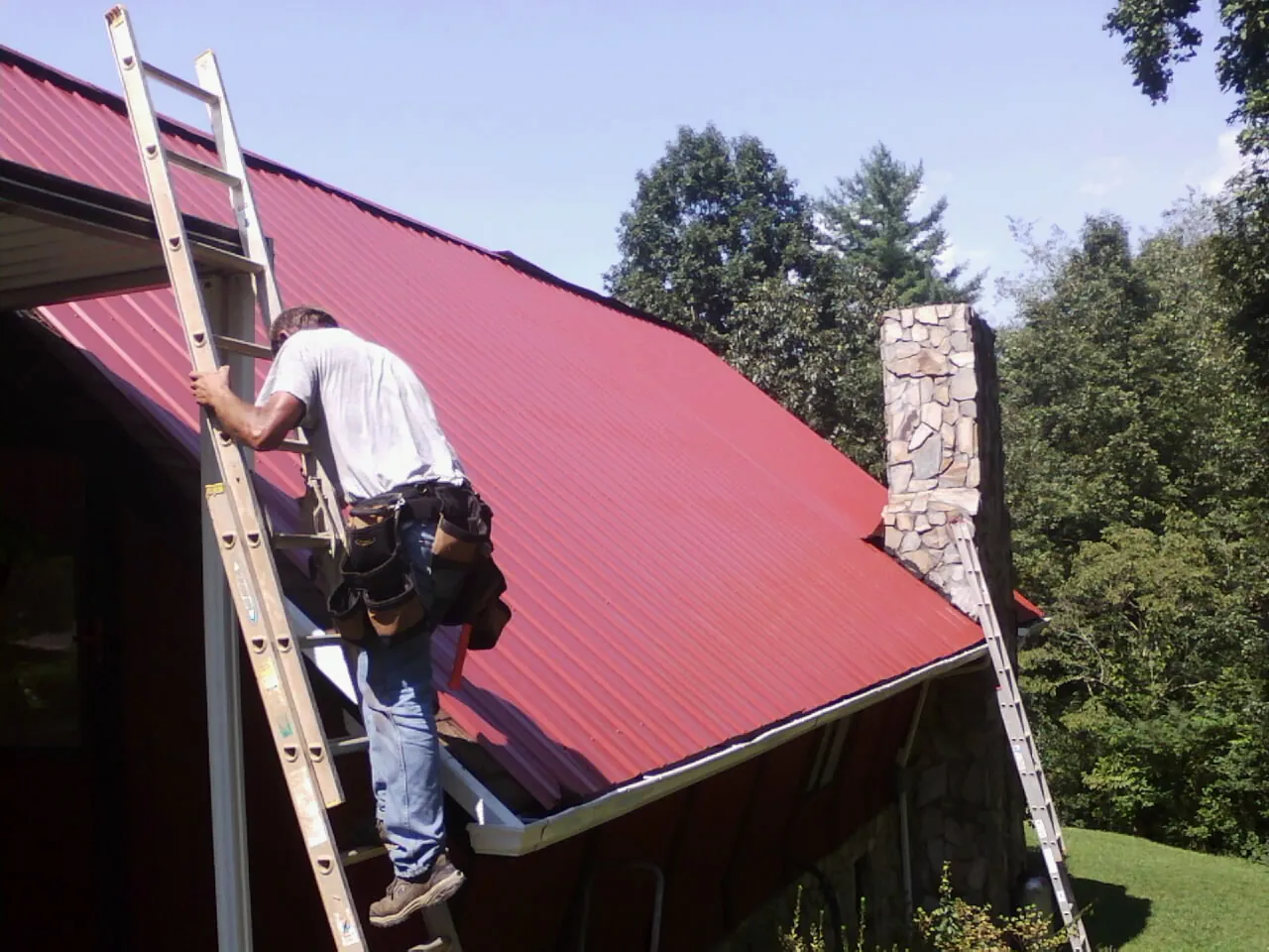 Roofer on a ladder beside a red metal roof and stone chimney — Soffit & Fascia Repair by Roofing Brothers in East Lansing